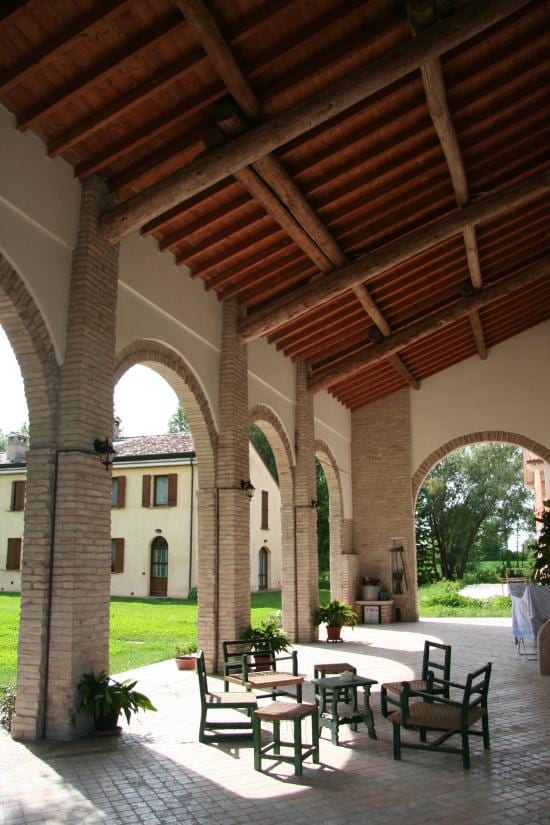 An airy covered patio features arched columns and a wooden beam ceiling. A set of four wooden chairs with woven seats surround a small table, while potted plants add a touch of greenery. A verdant lawn is visible beyond the arches.