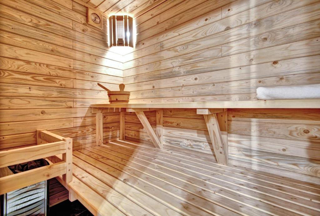 A wooden sauna interior features smooth, light-colored wood paneling and benches. Soft lighting is provided by a wall-mounted fixture, creating a warm ambiance. A wooden bucket and a rolled towel are neatly placed on a shelf, offering functional touches to the serene space.