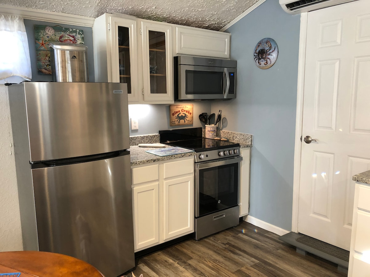 The kitchen features updated stainless steel appliances, including a refrigerator, microwave, and oven. Light-colored cabinetry complements the granite countertops. A small decorative piece on the wall adds a splash of color, while natural light illuminates the space through a nearby window.