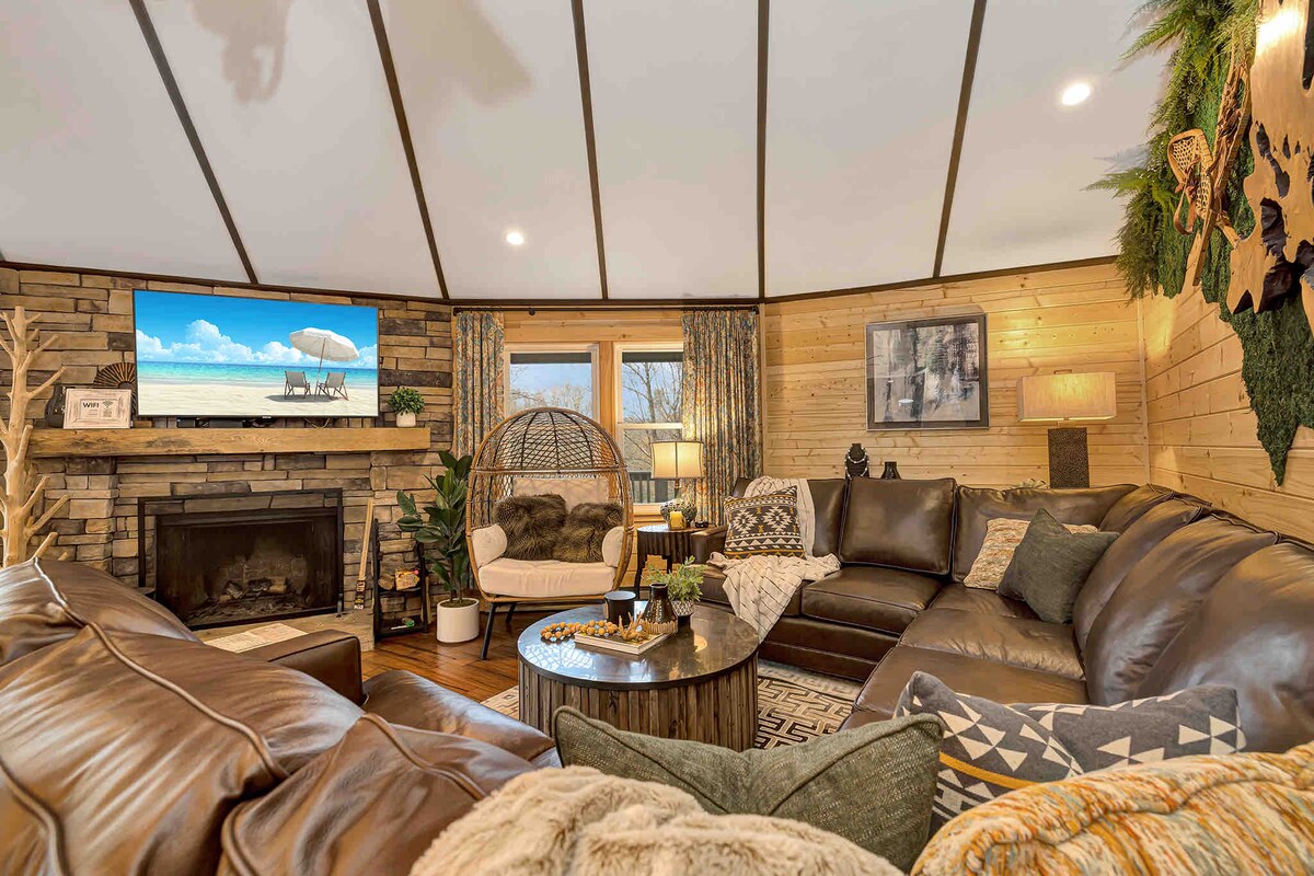The living area features a curved, dark leather sectional sofa surrounding a circular coffee table. A large flat-screen TV is mounted above a stone fireplace. Decorative accents include plants, cozy throws, and textured wall art, with natural light filling the space through large windows.