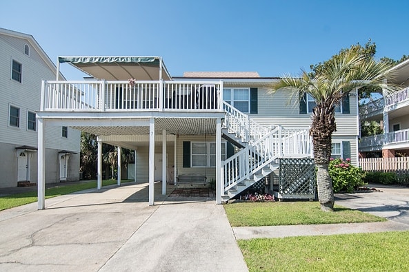 The exterior of a spacious two-story home is shown, featuring a large upper deck with an awning. A palm tree and well-maintained grass are visible in the yard, along with a concrete driveway leading to covered parking underneath the deck.
