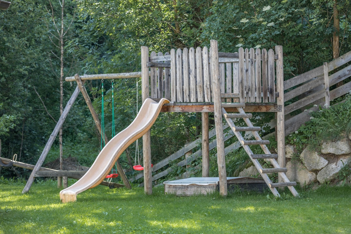 A wooden playground structure is visible, featuring a slide, climbing ladder, and swings. The structure is embraced by lush greenery, offering a playful environment for children. The grassy area surrounding the playground provides ample space for outdoor activities.