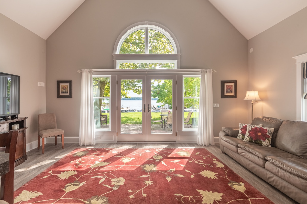 A spacious living area features large windows that provide views of Sodus Bay. Natural light fills the room, enhancing the warm tones of the area rug. A comfortable couch and a side chair are arranged around a coffee table, creating a welcoming space for relaxation.