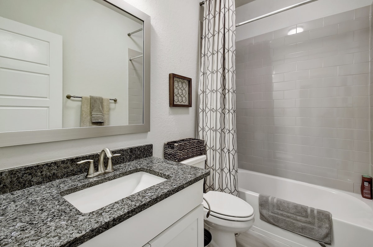 A modern bathroom features a white vanity with a sleek sink and granite countertop. A large mirror hangs above, reflecting the space. Gray tiles accent the shower area, which is lined with a textured curtain. A cozy towel is neatly placed nearby.