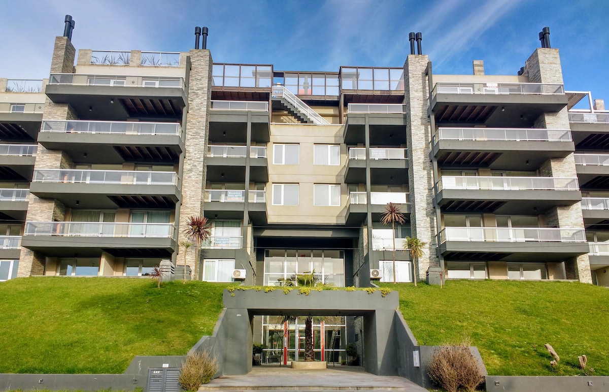 The modern building features multiple levels with a combination of stone and glass facades. Balconies extend from several units, offering outdoor space. Lush greenery surrounds the entrance, which is highlighted by a clear glass door and large windows that reflect natural light.
