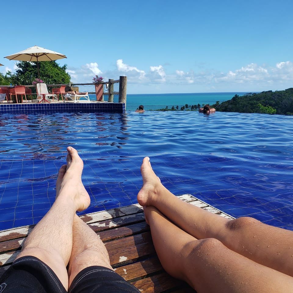 An infinity pool is shown, with clear blue water blending into the horizon. Two pairs of legs are visible at the edge, with a wooden deck beneath. In the background, lounge chairs and an umbrella provide shade, while distant figures are seen enjoying the water.