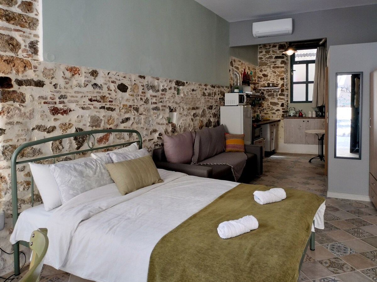 A spacious studio apartment features a double bed with soft linens and decorative pillows. A sofa-bed and kitchenette are visible in the background, surrounded by rustic stone walls. Natural light filters through a window, enhancing the warm and relaxing ambiance of the space.
