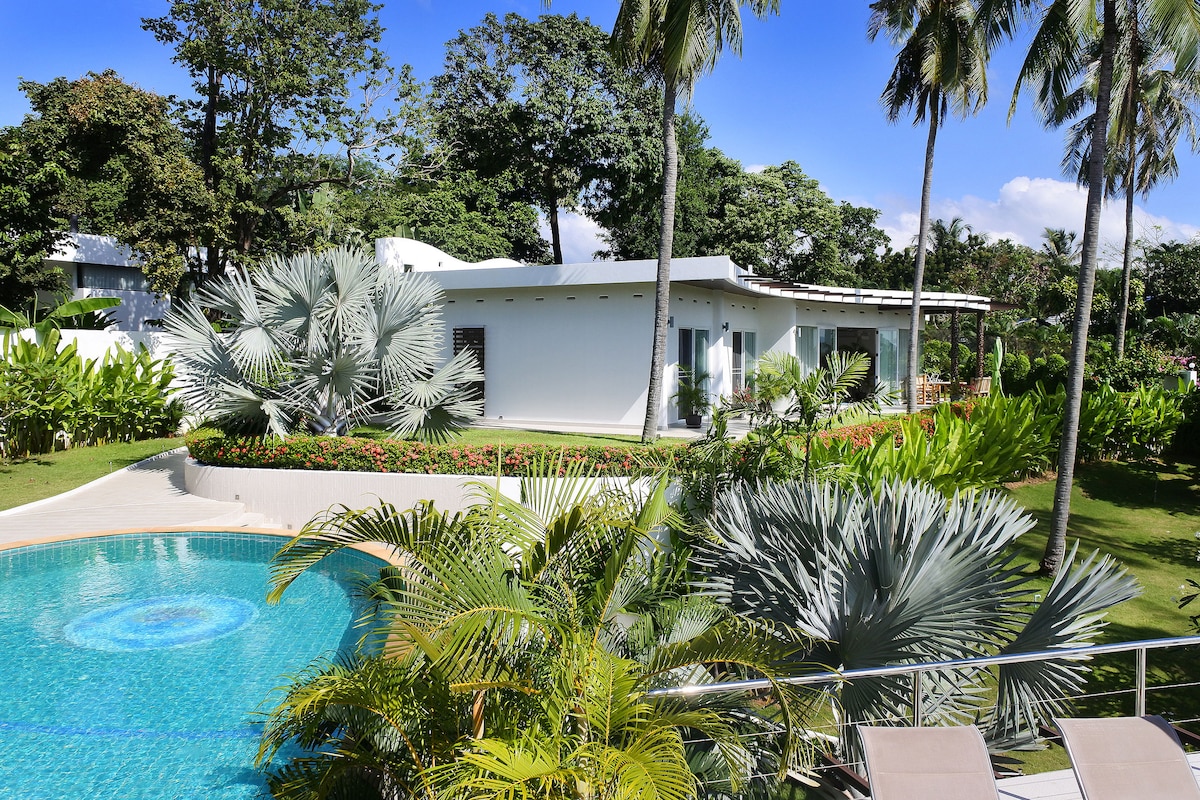 A modern villa is set against a backdrop of lush tropical greenery, with a shimmering swimming pool in the foreground. Palms and various plants surround the property, emphasizing the tranquil and spacious outdoor area designed for relaxation.