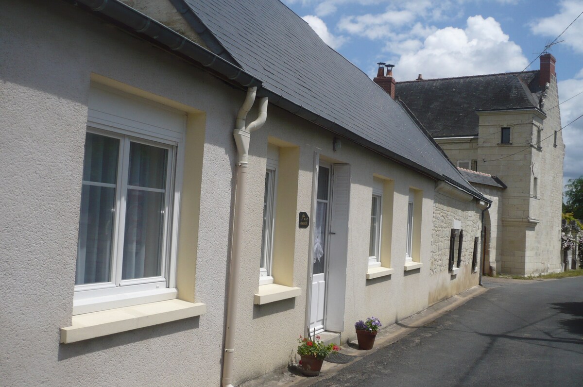 A single-story house features a light-colored exterior with multiple windows framed in white. Potted flowers adorn the entrance, adding a touch of color. The quiet street is lined with a mix of architectural styles, showcasing the charm of the surrounding area.