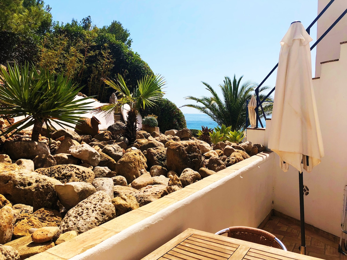 A relaxing patio area is shown, featuring a wooden table and a pair of lounge chairs surrounded by a variety of plants and rocks. A partial view of the sea is visible beyond the greenery, complemented by clear blue skies.