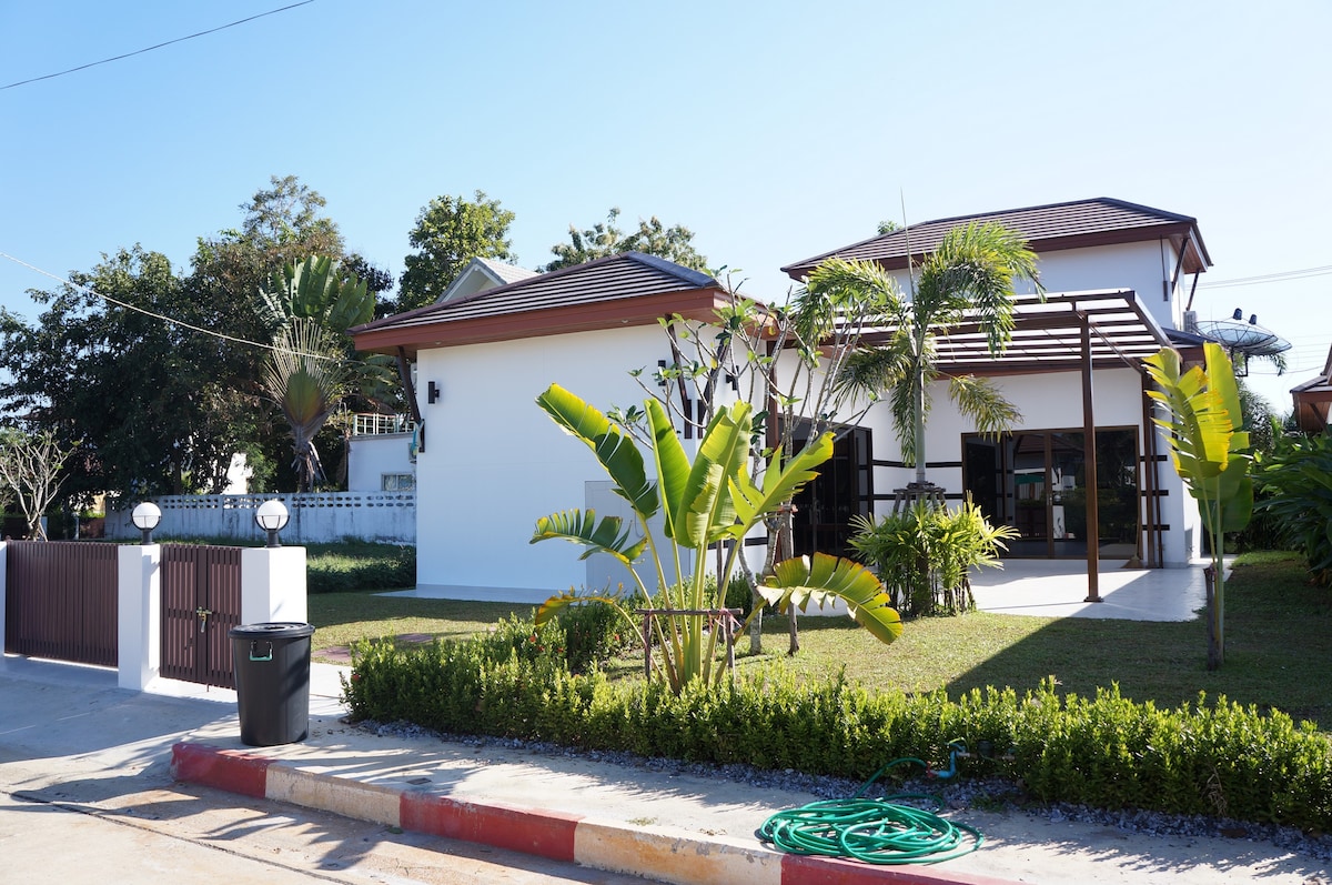 A modern two-story house is presented, featuring a white exterior and brown accents. Tropical plants and palm trees surround the entrance, with a well-maintained garden leading up to the porch area. A spacious driveway is visible, framed by a decorative fence.
