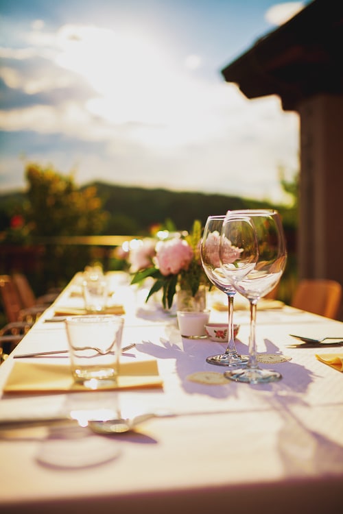 A dining table is set outdoors, featuring a white tablecloth and neatly arranged utensils. Clear glasses are placed alongside small decorative plants. Soft sunlight illuminates the scene, and distant hills can be seen through the background foliage.
