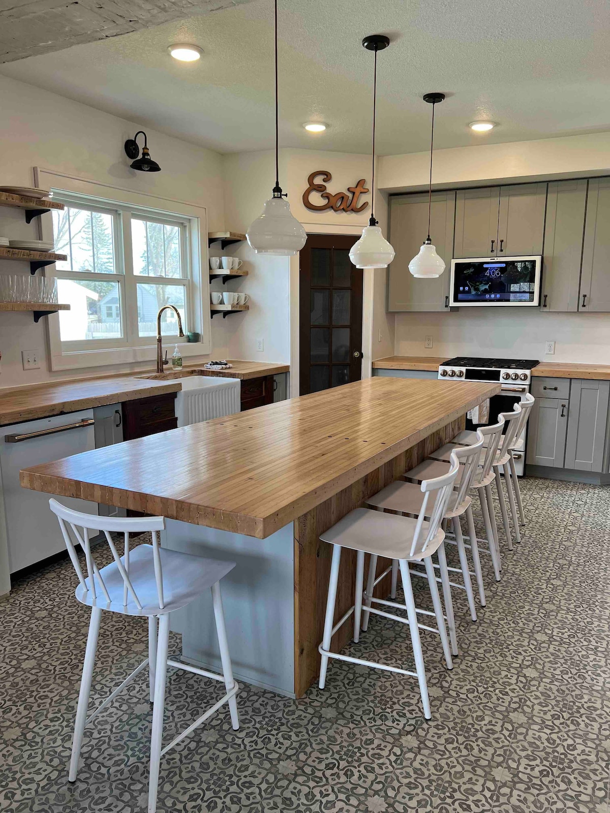 An inviting kitchen features a long wooden island with white bar stools, providing ample seating. Modern cabinetry and a microwave are present, while pendant lights hang above, enhancing brightness. Large windows allow natural light to enter, and decorative elements add a touch of charm.