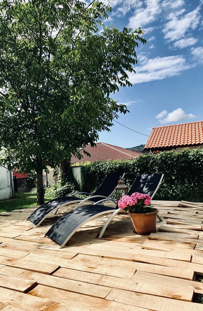 A serene outdoor area is featured, with two lounge chairs positioned on a wooden deck. A potted plant with vibrant pink flowers adds a colorful touch, while a tree provides shade. The clear blue sky and gentle clouds create a peaceful setting.