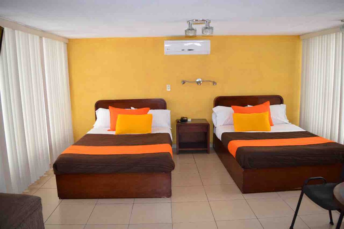A bright room features two spacious beds with brown and orange bedding. Cushions in yellow add a pop of color. A small nightstand with a lamp is visible beside the beds. Large windows covered with sheer curtains allow soft natural light to fill the space.