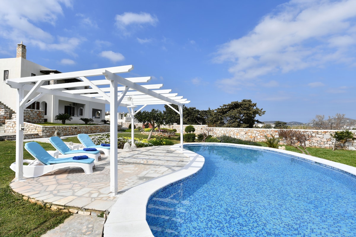 A curved pool is surrounded by a stone patio and lush greenery, with white lounge chairs positioned under a pergola. The serene outdoor space offers a tranquil setting, complemented by bright blue skies and scattered clouds in the background.