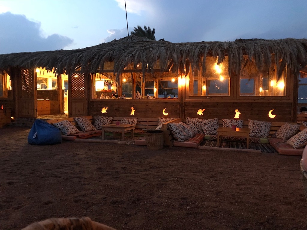 A beachside lounge area is featured, with thatched roofs and warm lighting creating a relaxed atmosphere. Plush seating with patterned cushions sits on sandy ground, while large bean bags offer additional comfort. Glowing decorations are visible across the wooden structure, enhancing the tranquil setting.