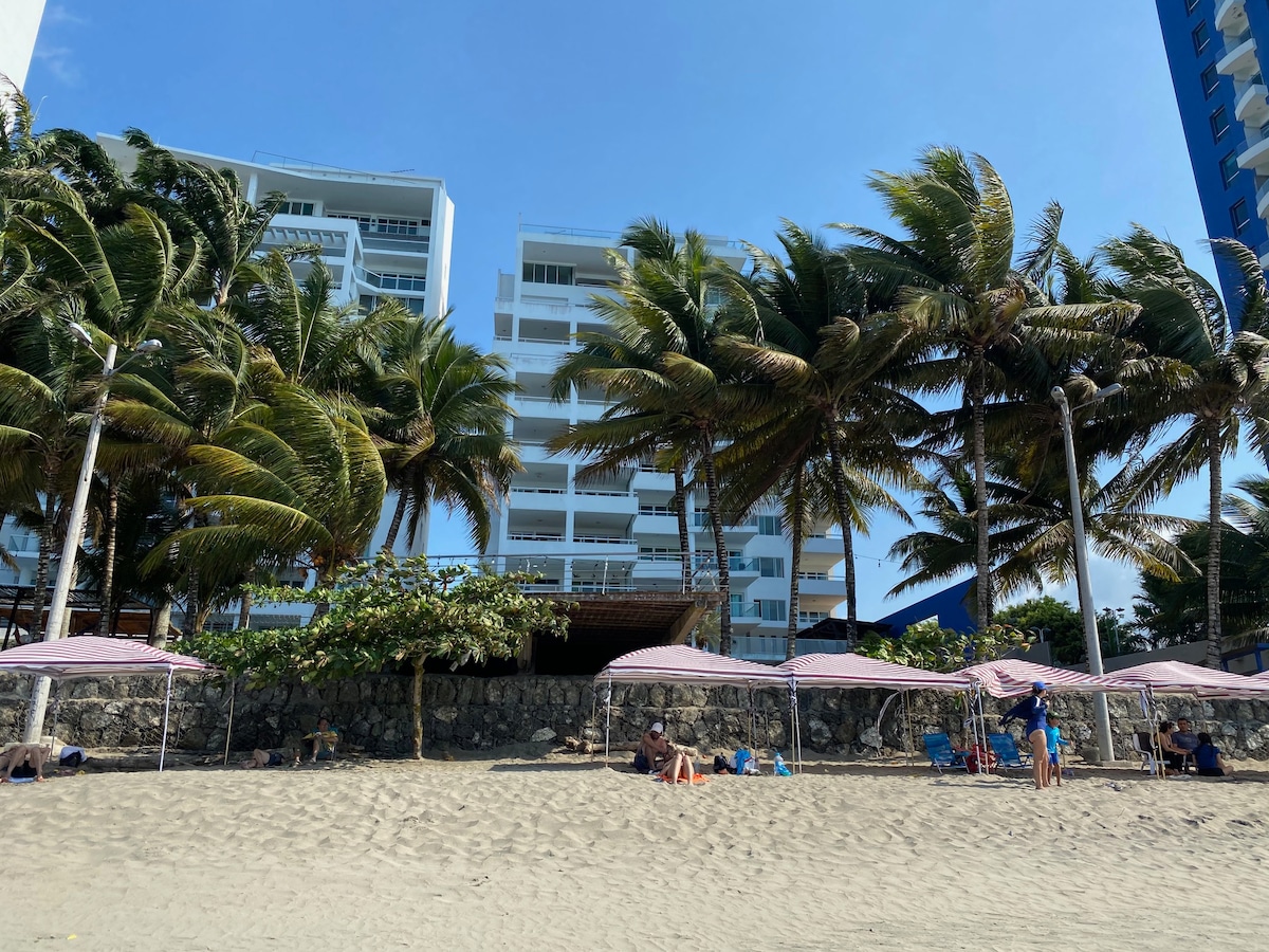 The image captures a sandy beach scene, with tall palm trees swaying in the breeze. In the background, a modern multi-story building is visible. Several beach canopies provide shade for guests enjoying the outdoor space, with people scattered along the shoreline.