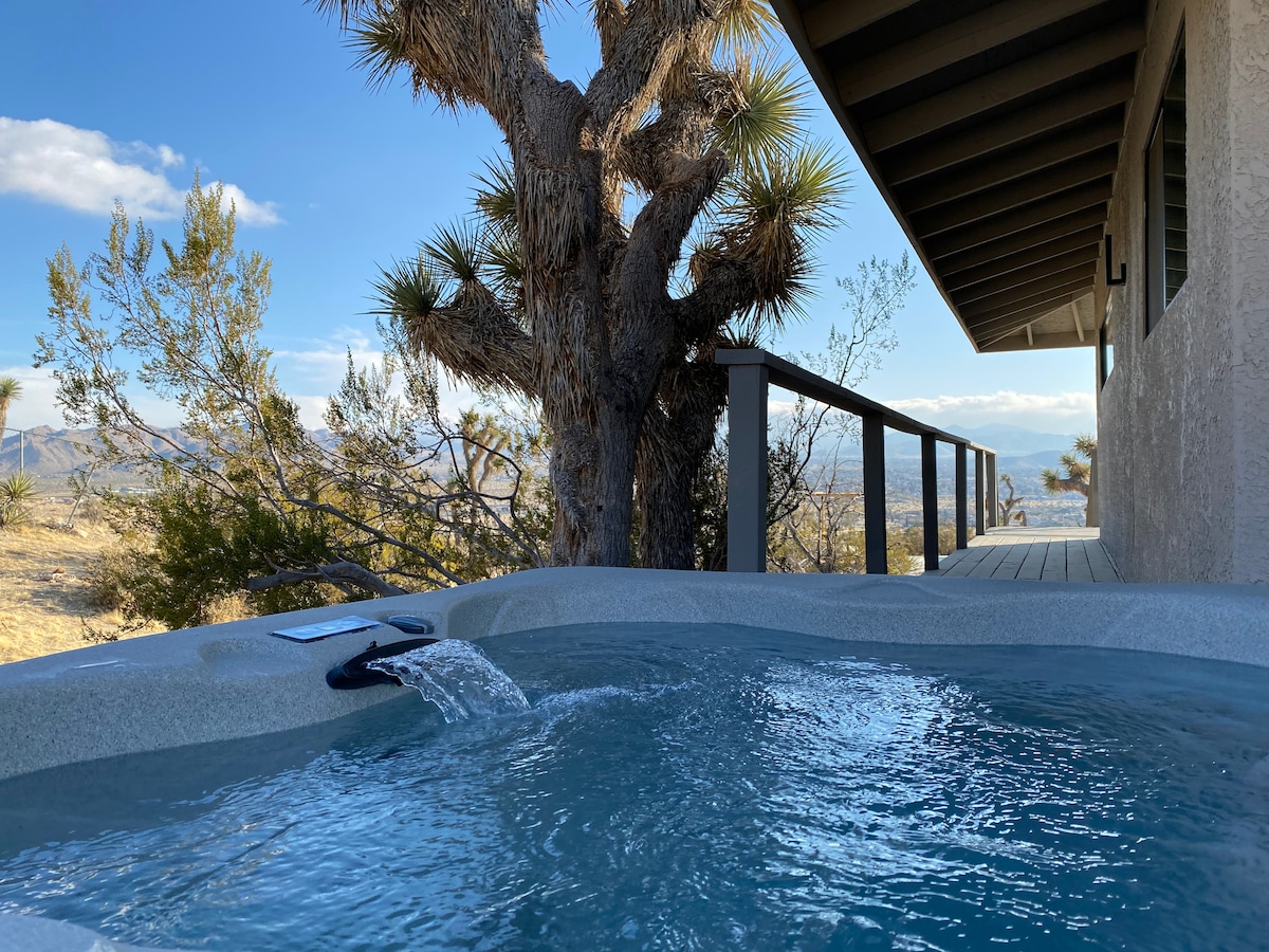 A hot tub is positioned on a deck with a view of the surrounding landscape. A nearby Joshua Tree and the house can be seen in the background, with clear skies and distant hills contributing to the serene atmosphere.