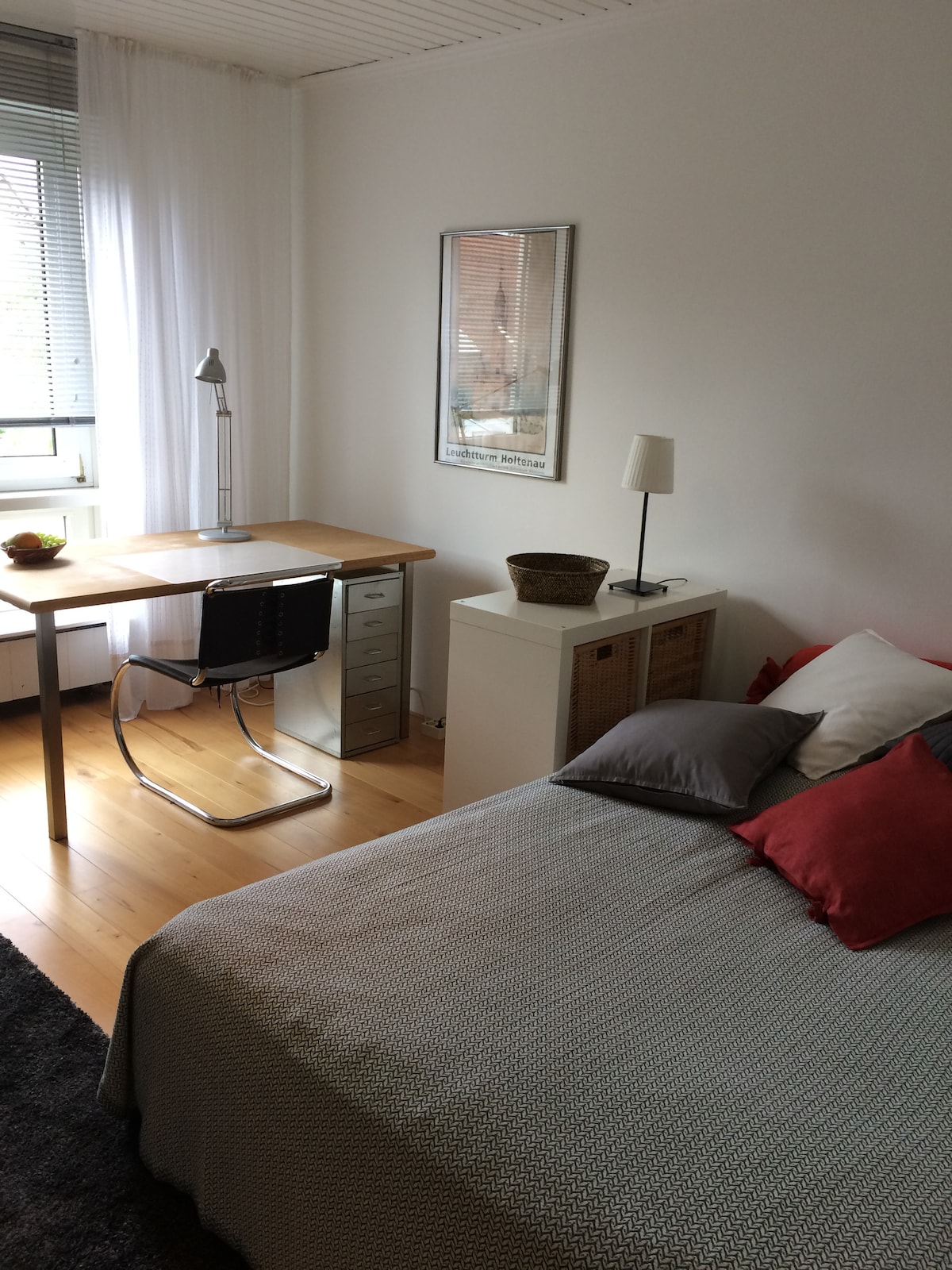 A quiet bedroom is equipped with a spacious bed adorned with colorful pillows. A wooden desk with a lamp and a chair is positioned nearby, creating a functional workspace. Natural light enters through a window, and warm wooden flooring complements the soft tones of the decor.