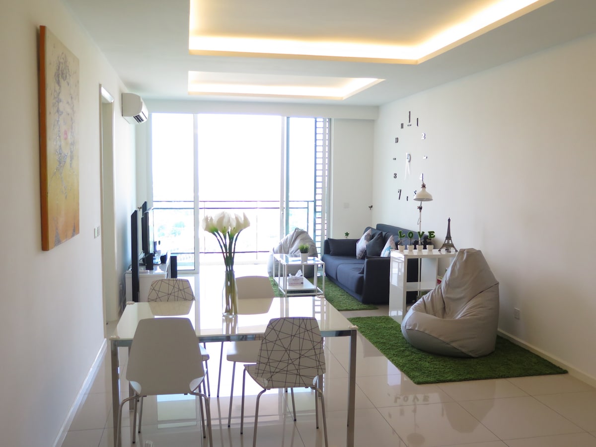 A modern living space is depicted with a sleek dining table and white chairs. A plush couch is positioned beside a large window offering city views. Decorative elements include a small plant, a bean bag, and wall art, creating an inviting atmosphere.