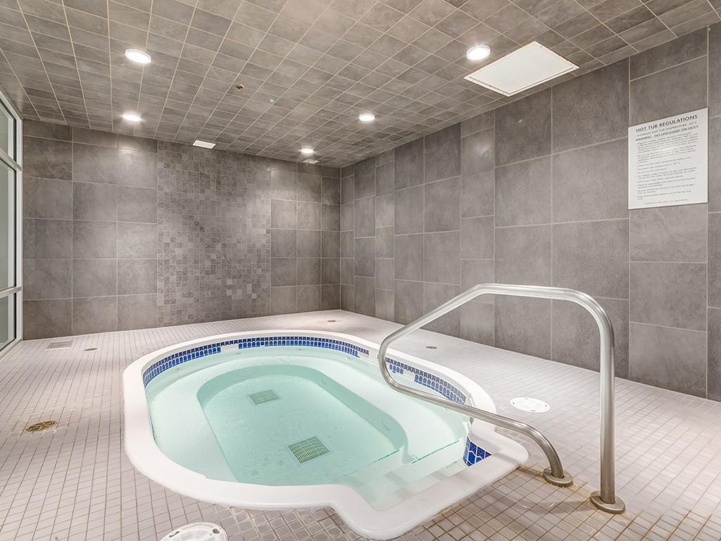The indoor hot tub is set against a backdrop of gray tiled walls and floor. Soft lighting creates a calming ambiance, and a handrail is positioned for accessibility. The tub features blue tile accents along the interior perimeter, promoting a serene and relaxing environment.