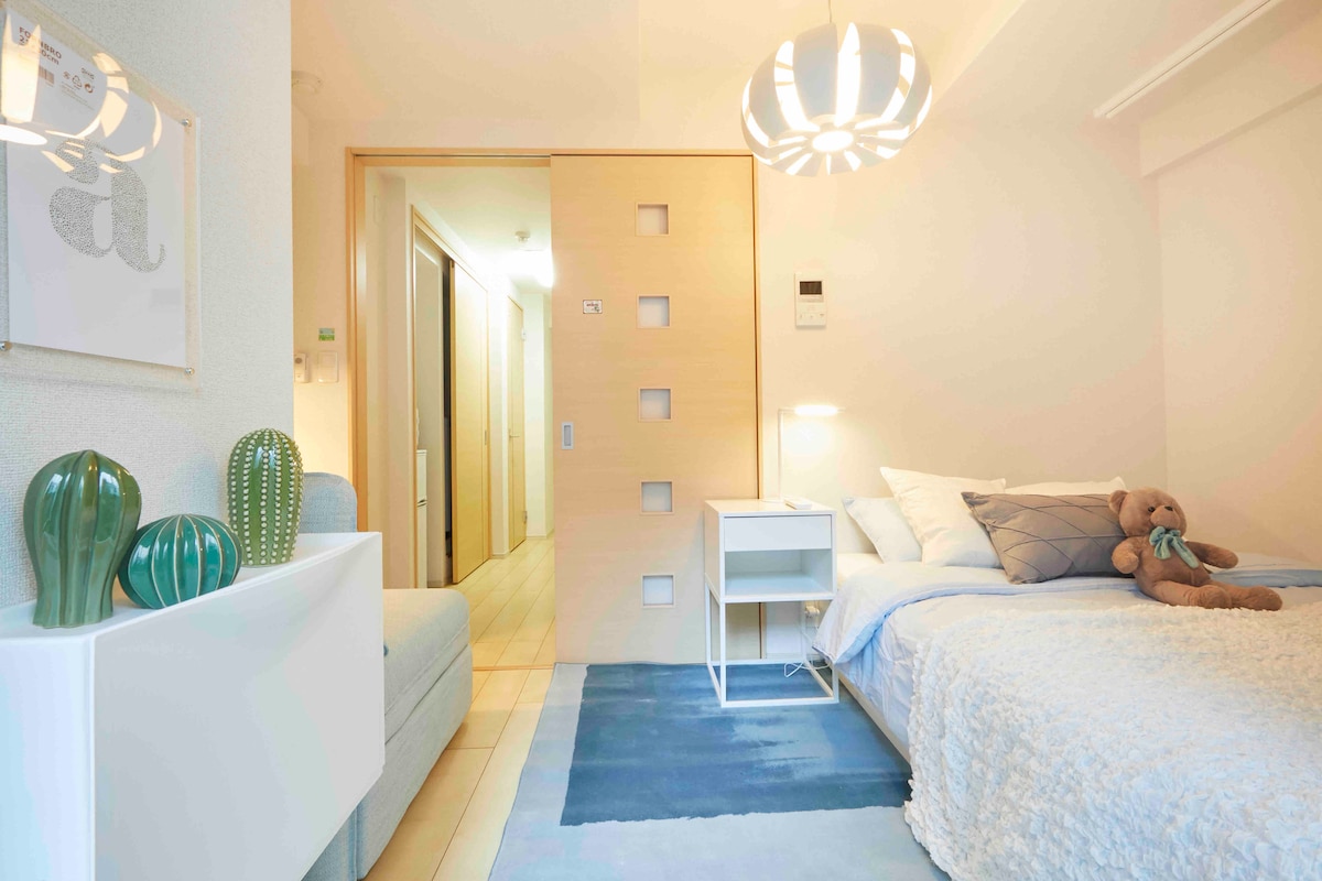 A comfortable sleeping area is presented with a neatly made bed and soft bedding. A bedside table holds a decorative lamp. A plush teddy bear is positioned on the bed, while a decorative rug adds a touch of color to the floor. Natural light enters through a nearby opening.
