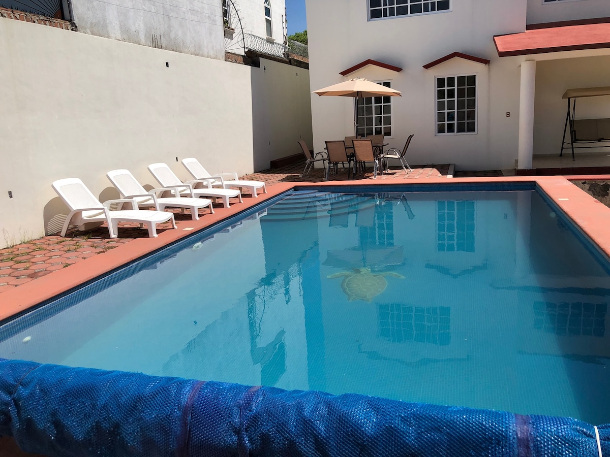 A private pool area is seen featuring a clean, blue swimming pool surrounded by a tiled deck. Several lounge chairs are positioned nearby, alongside an umbrella providing shade. The backdrop includes a two-story building with large windows allowing natural light.