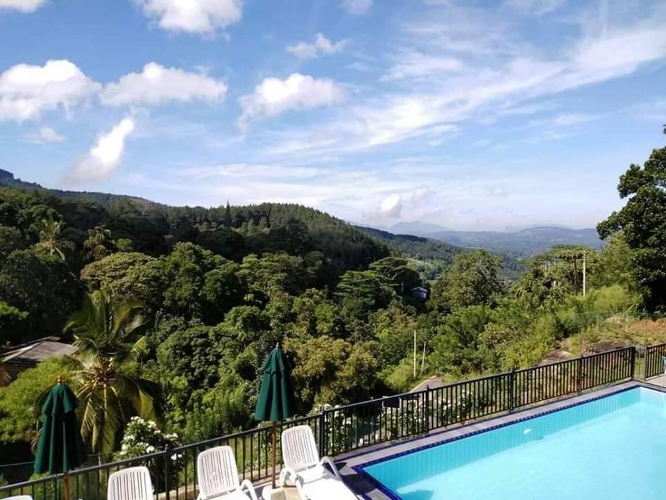 An outdoor swimming pool is visible, surrounded by sun loungers. Lush greenery and distant hills create a serene backdrop under a clear blue sky, with a few clouds scattered above. The area is enhanced by the presence of umbrellas providing shade.