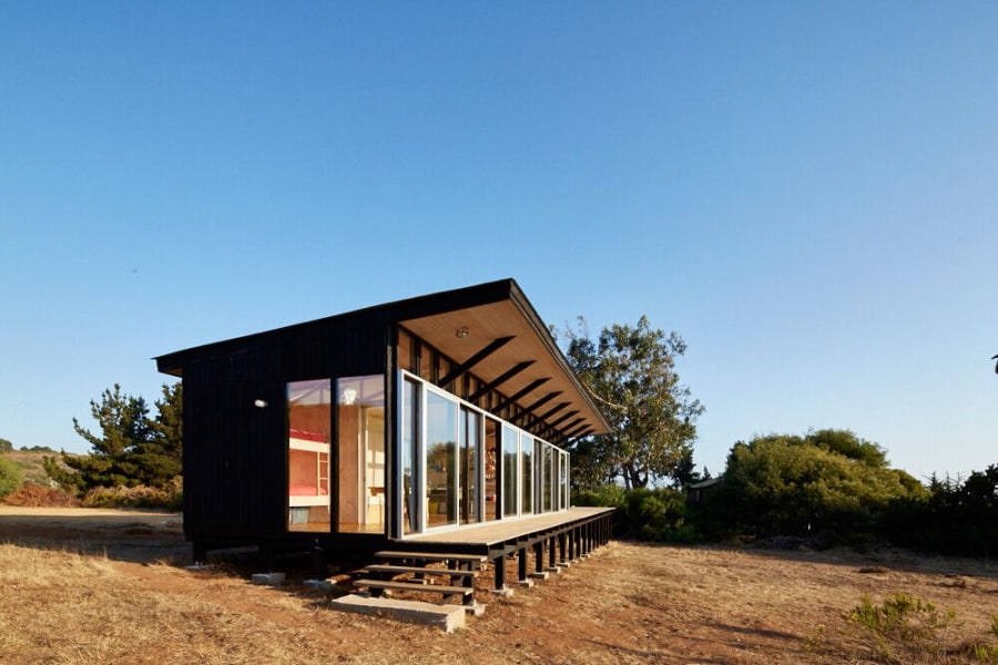 A modern ecological house is presented, featuring a striking black exterior and expansive glass windows. The structure is elevated above the ground, offering a view of the surrounding landscape and nature. The clear blue sky completes the serene setting.