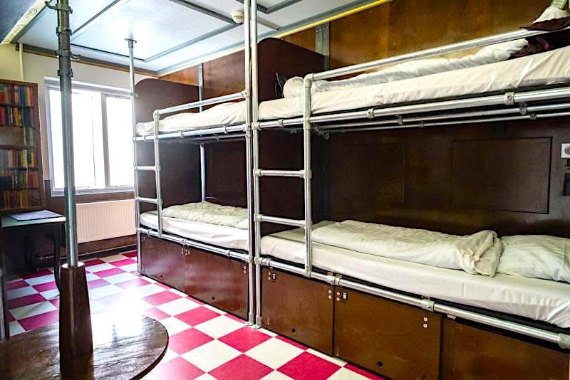 An interior view of a dormitory room features two sets of metal bunk beds lined against a wall. The floor is decorated with a red and white checkered pattern. A window allows natural light to brighten the room, which is complemented by wooden paneling.