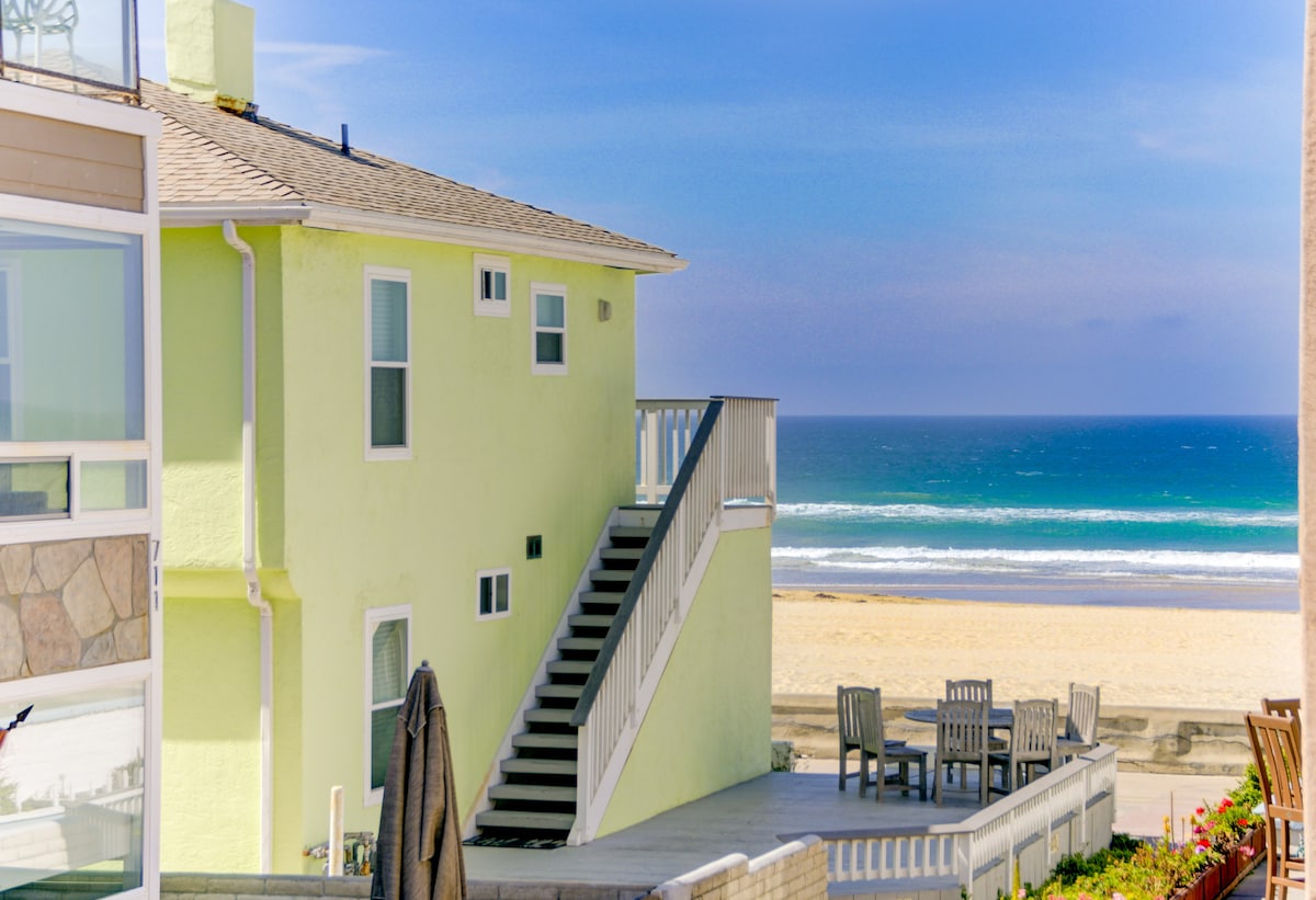 A vibrant green beach house is positioned steps from the sandy shore. A multi-level structure features a staircase leading to an upper level, with panoramic ocean views visible from a nearby outdoor dining area. The serene beachfront offers a wide expanse of sand and gentle waves.