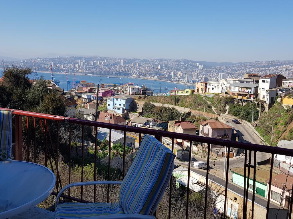 A scenic view from a balcony showcases a sprawling coastal landscape, including a picturesque harbor and vibrant cityscape. Colorful houses are visible on the hillside, while the serene blue water reflects the clear sky. Comfortable striped chairs are positioned around a small table, inviting relaxation.