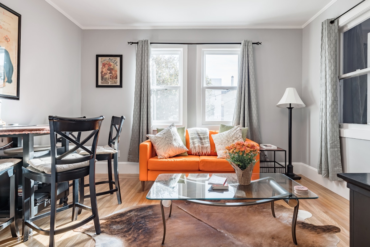 The living room features a bright orange couch complemented by natural light from two windows. A glass coffee table is centered on a textured area rug, while a standing lamp and decorative vase with flowers add warmth to the space. Seating includes four black chairs at the dining area.