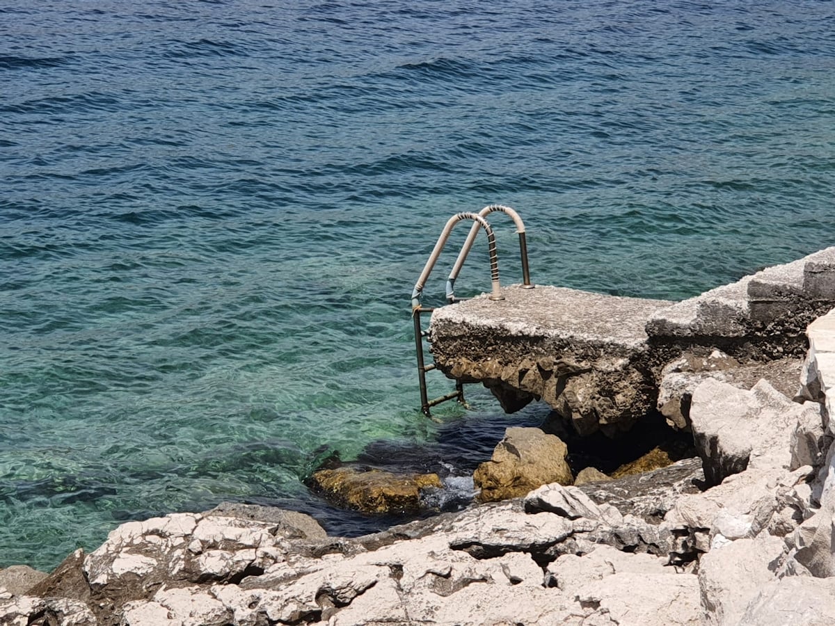 A concrete platform extends over the clear, blue-green water, featuring a metal ladder for access. Rocky ledges border the platform, providing a natural coastal view. The gentle waves can be seen lapping against the stones, creating a serene seaside environment.