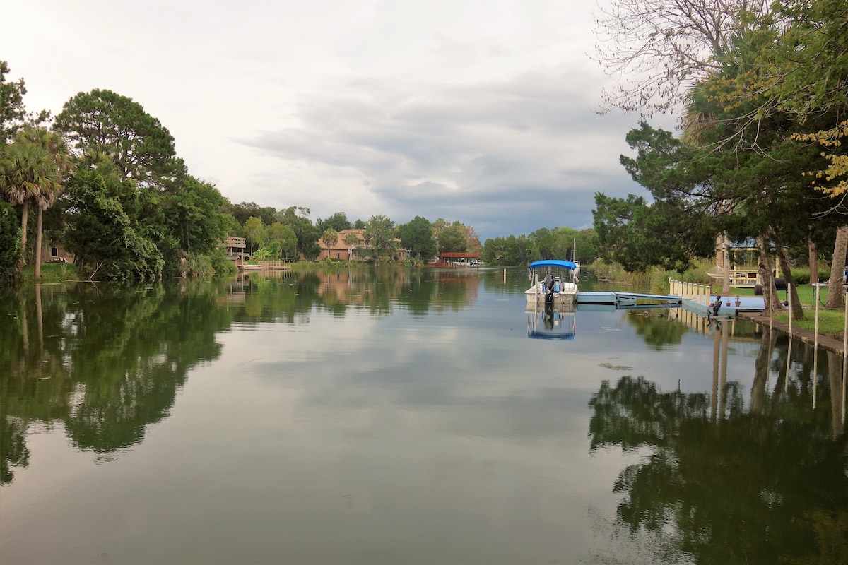 A calm lagoon is depicted, featuring still waters that reflect the surrounding greenery and wooden structures. A small boat is docked nearby, adding to the serene atmosphere. The sky shows varying shades of grey, hinting at a peaceful moment in nature.