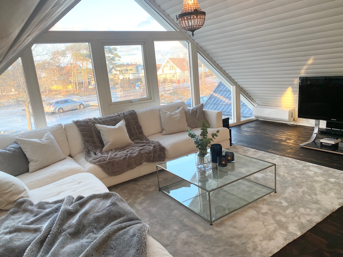 A spacious living room features a large sectional sofa adorned with soft pillows and a warm throw. A glass coffee table sits on a light gray area rug. Large windows allow natural light to fill the room, providing views of the surroundings.