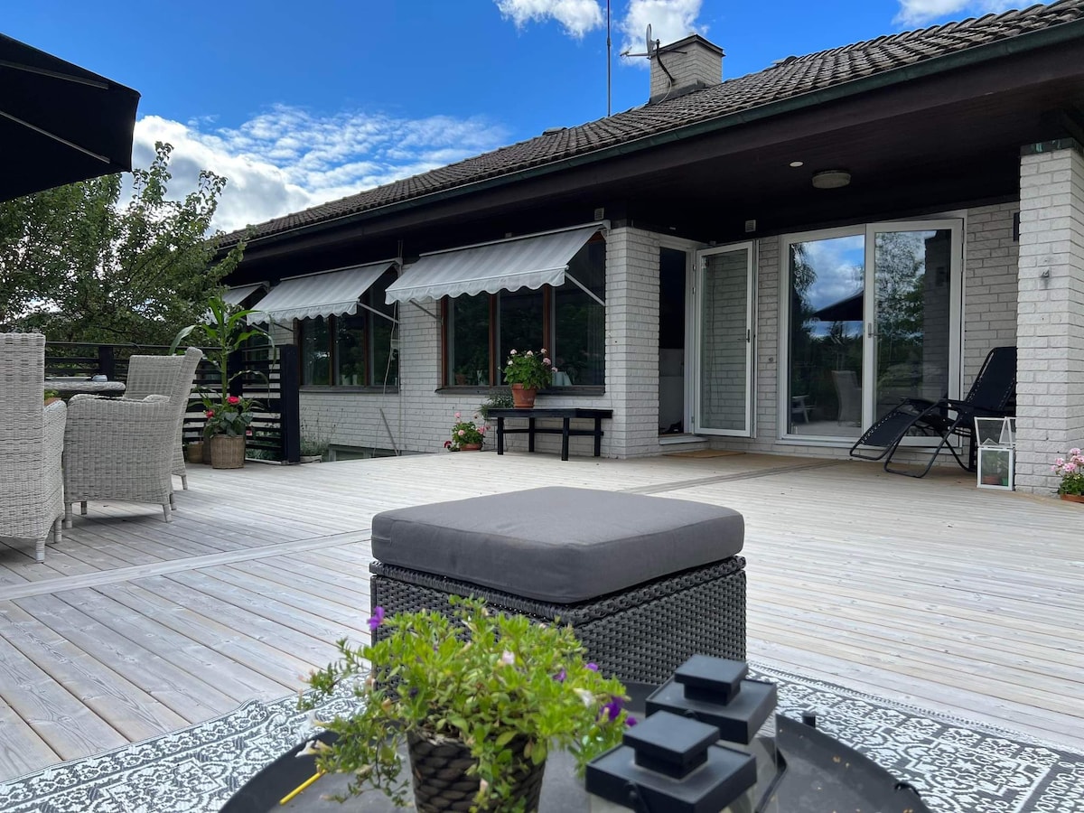 A spacious outdoor deck is featured, surrounded by greenery and adorned with potted plants. Comfortable seating arrangements are visible, alongside a low table topped with decorative items. Large windows lead into the interior, allowing natural light to brighten the area.
