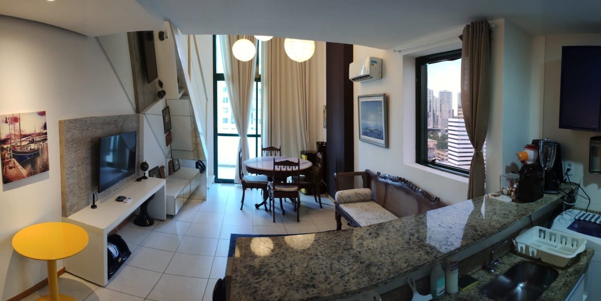 A modern open-concept living area showcases a well-appointed kitchen with a sleek granite countertop. A comfortable seating arrangement, including a vintage sofa and dining table, creates a welcoming environment. Large windows let in natural light, offering views of the cityscape beyond.