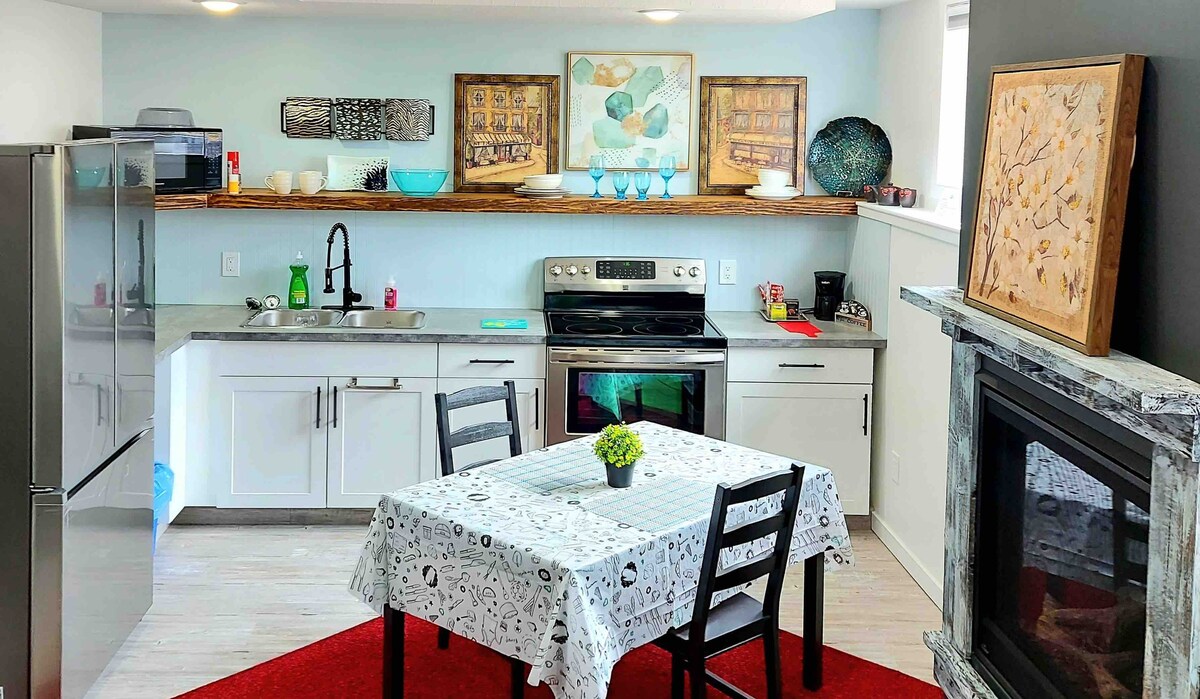 A modern kitchen is showcased with stainless steel appliances, including a refrigerator and stove. An island counter sits beside a dining table set for four, covered with a patterned tablecloth. Natural light brightens the space, complemented by decorative wall art and a small green plant.