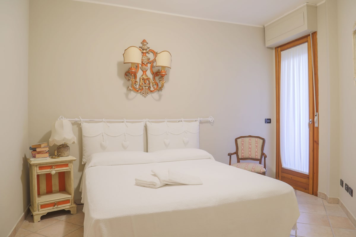 A well-appointed bedroom features a spacious bed with white bedding, complemented by a vintage chair and a nightstand. A decorative wall sconce adds charm, while a window provides natural light, with light curtains softly diffusing the view.