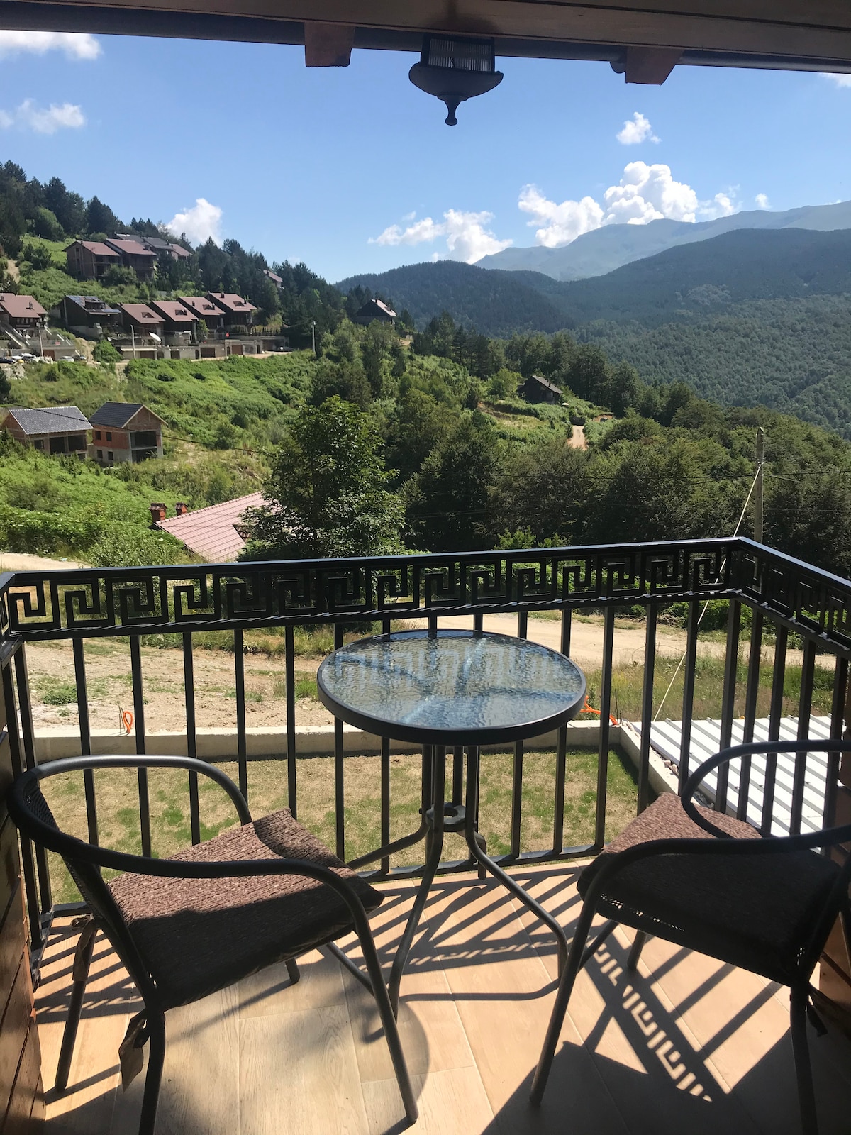 A private balcony features two chairs and a small table, overlooking lush green hills and distant mountains. Scenic views of the surrounding landscape are visible, highlighting the natural beauty of the area and providing a tranquil space for relaxation.