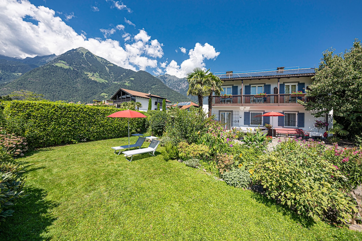 A sunlit garden is surrounded by lush greenery and colorful flowers, featuring two lounge chairs beneath red umbrellas. The backdrop showcases a charming multi-story building with balconies. Majestic mountains and a clear blue sky complete the serene outdoor environment.