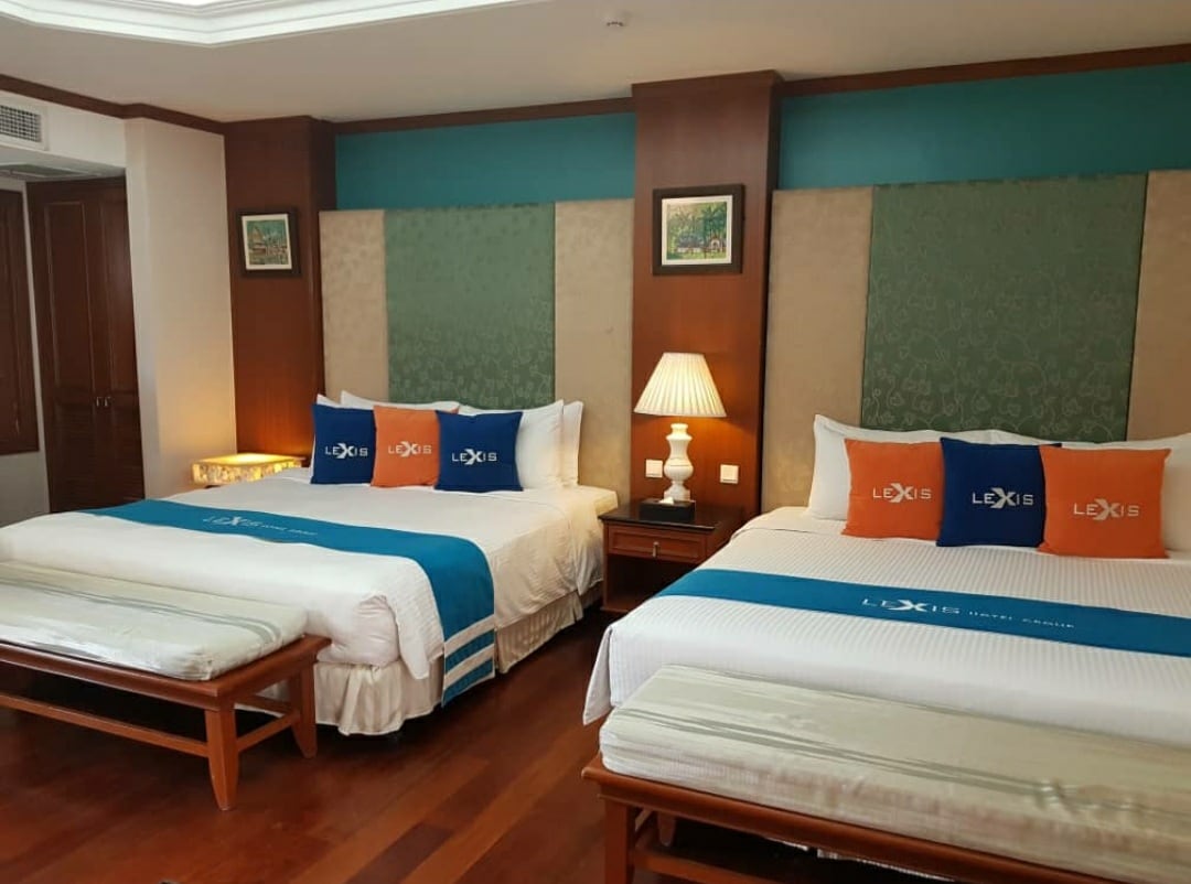 A spacious guest room is equipped with two large beds, each adorned with bright blue and orange accent pillows. Soft lighting is provided by a bedside lamp, and wooden furniture adds a warm touch to the space. Textured wall panels create visual interest behind the beds.