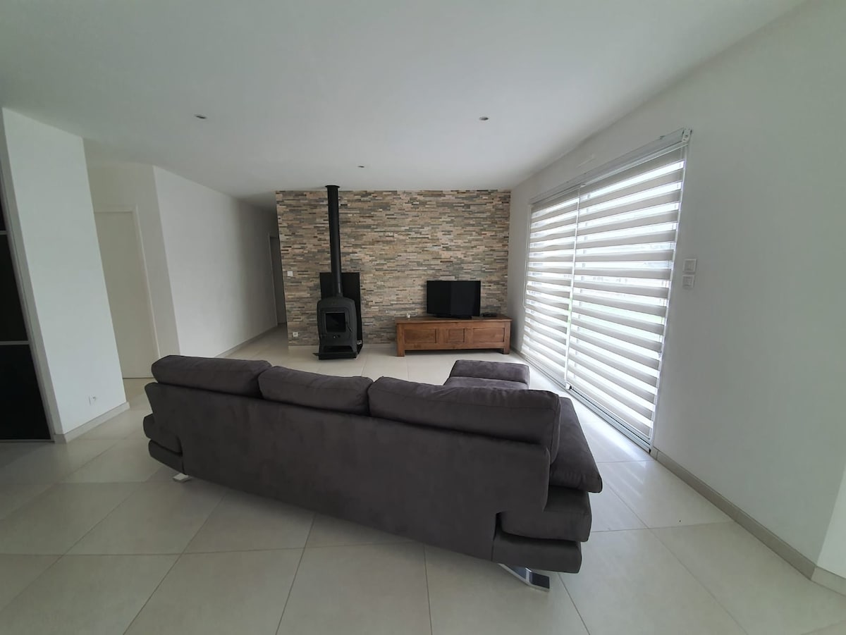 The spacious living area is characterized by a modern layout, featuring a comfortable sectional sofa facing a wooden entertainment unit and a flat-screen TV. A stone wall adds texture, while large windows with horizontal blinds allow natural light to fill the space.