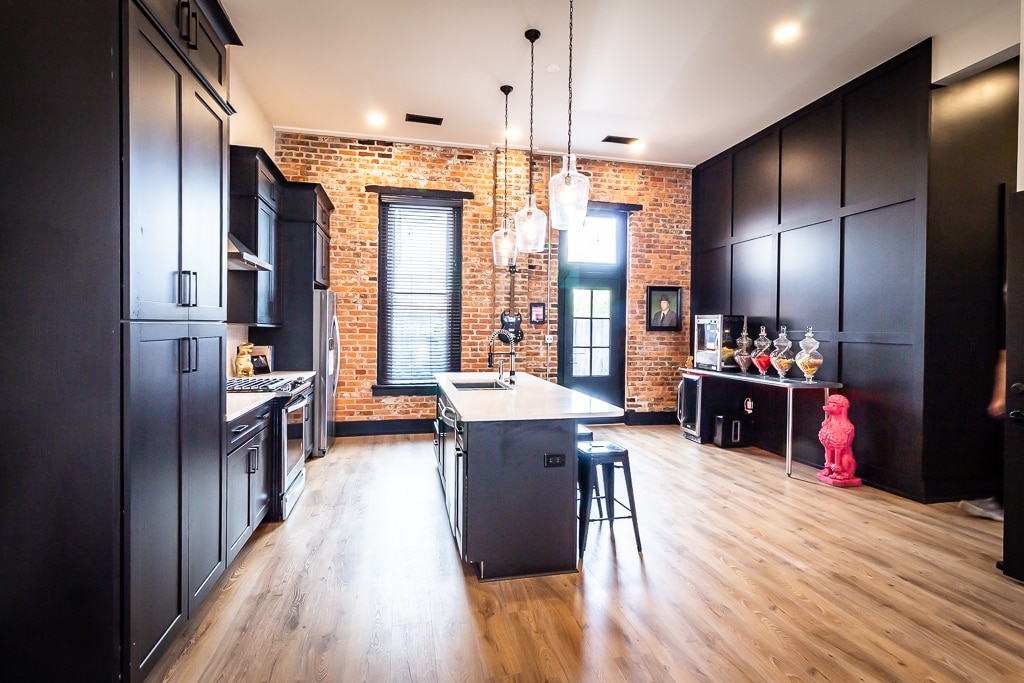 A modern kitchen showcases sleek black cabinetry and a spacious island with seating. Exposed brick walls add character, while pendant lighting provides illumination. A bright, open space features large windows and hardwood flooring, completing a contemporary ambiance.