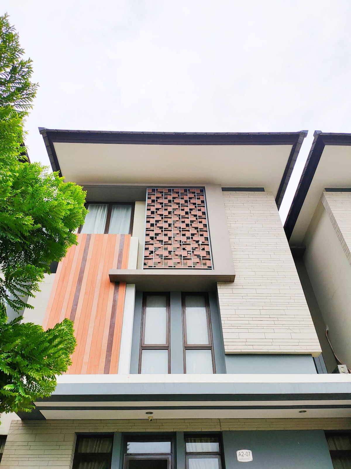 A modern two-story house is presented, featuring a combination of textured panels and large windows. Geometric patterns decorate the upper section, complemented by natural wood accents. Lush green foliage lines the front, contributing to a serene appearance against a cloudy sky.