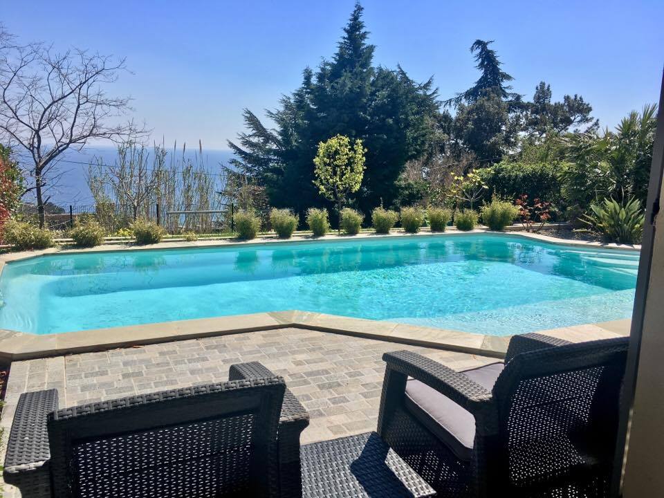 A private pool is set in a landscaped garden, surrounded by lush greenery. Two comfortable chairs are positioned at the edge of the pool, providing a serene view of the water and distant landscape under a clear blue sky.