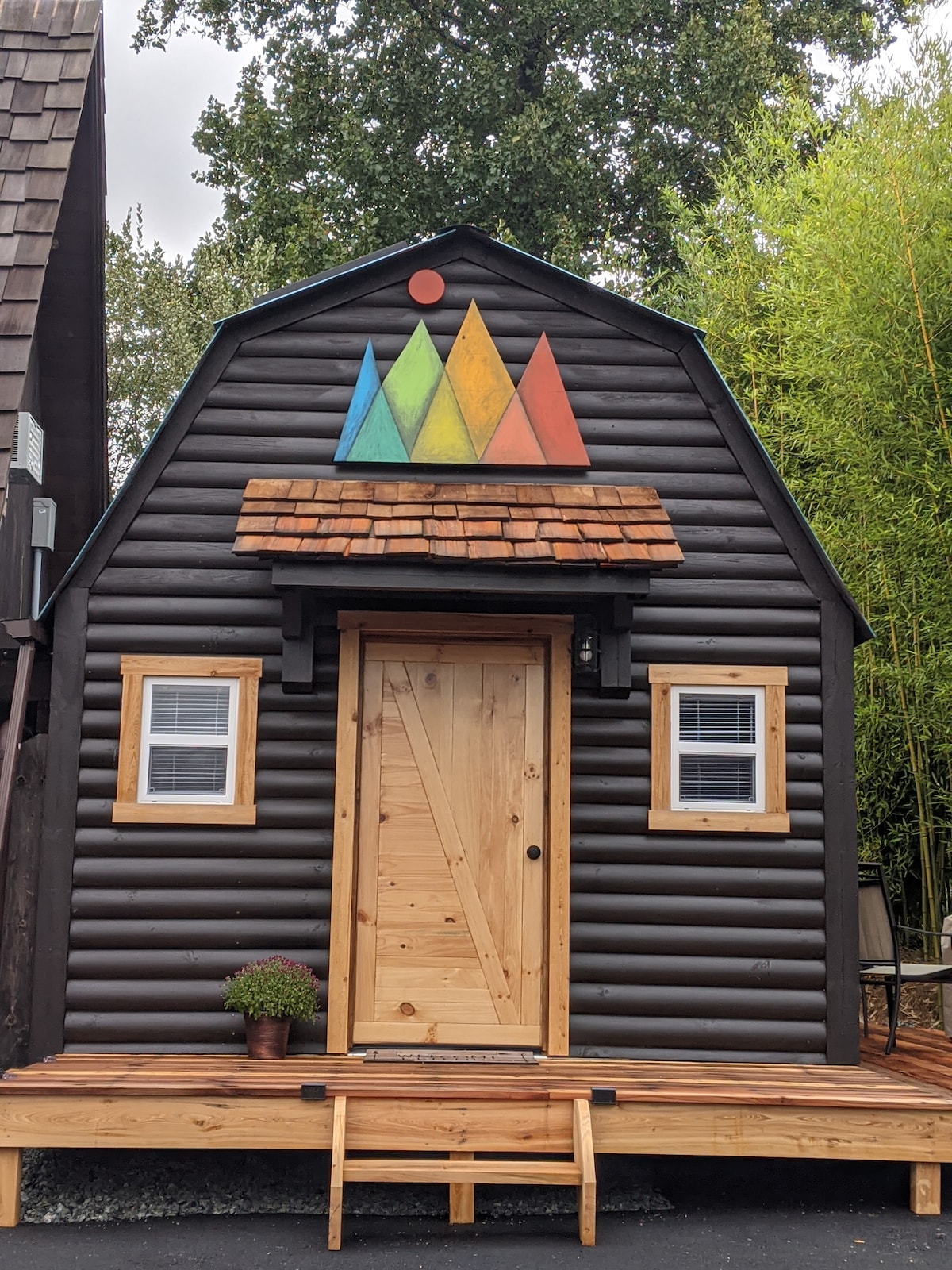 The exterior showcases a charming small structure with a black wooden façade, accented by a colorful triangular mountain design above the door. A welcoming wooden door is framed by symmetrical windows, with a small potted plant positioned at the entrance on a wooden porch.