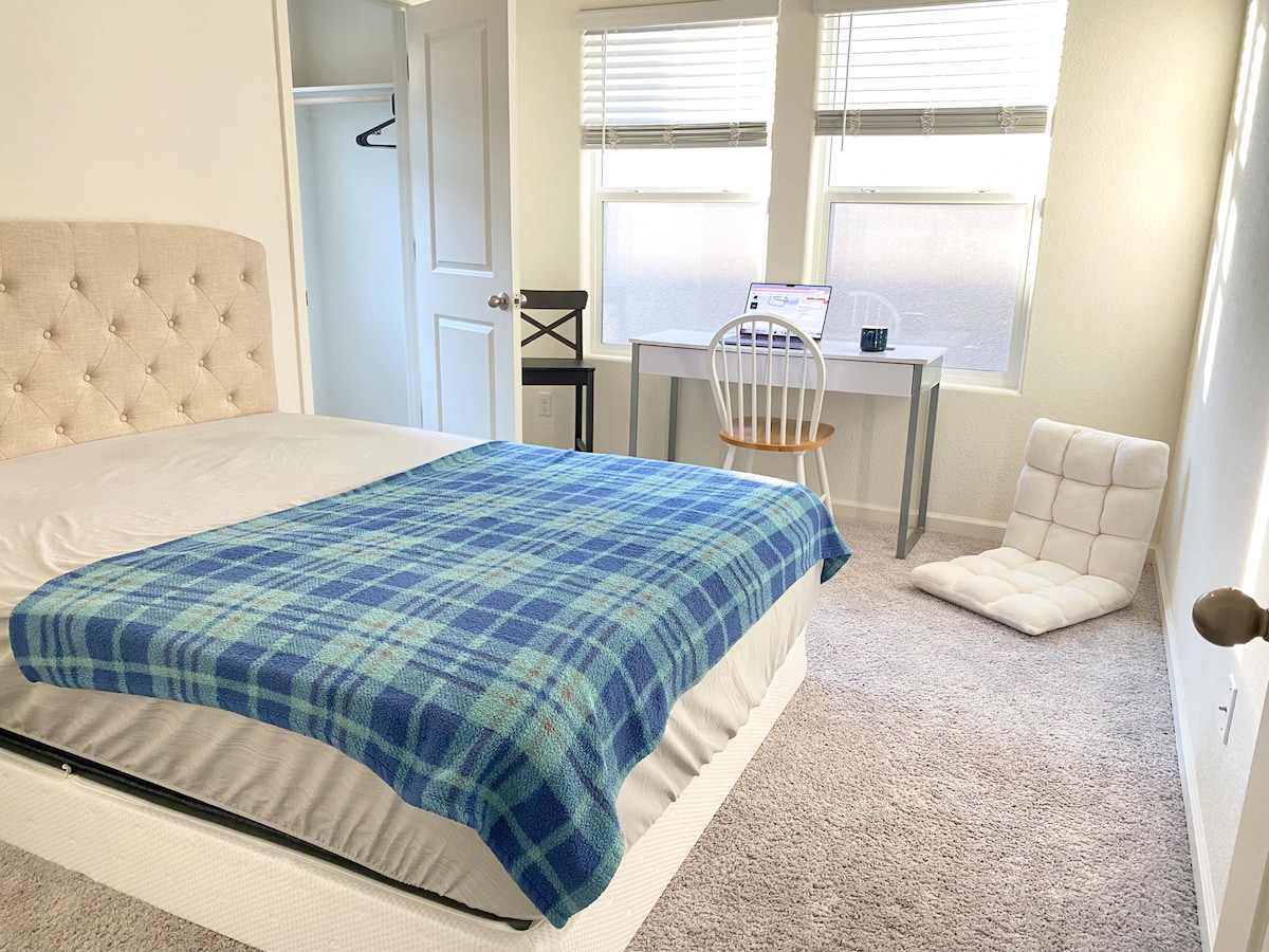 A well-appointed bedroom features a spacious bed adorned with a plaid throw. Natural light is provided by two windows, while a small desk with a chair is positioned nearby. A cushioned floor chair adds additional seating to the space.